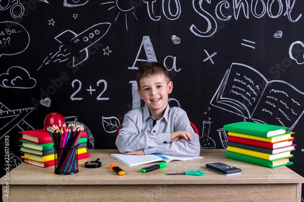 Obraz Happily smiling excellent schoolboy sitting on the desk with books, school supplies, with both arms leaned one to another