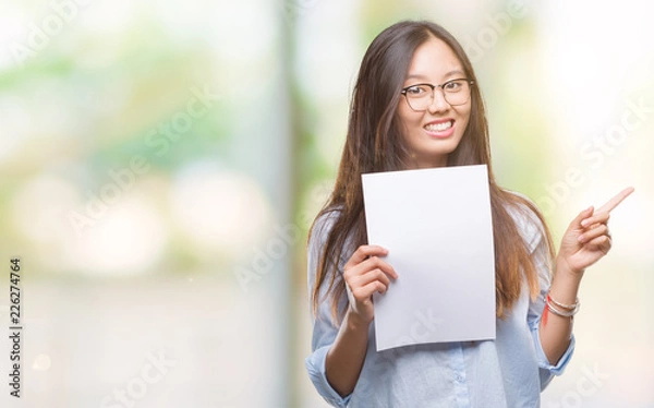 Fototapeta Young asian woman holding blank paper over isolated background very happy pointing with hand and finger to the side
