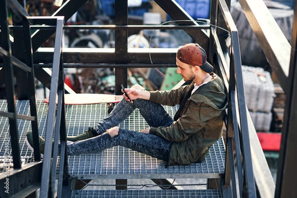 Obraz Young handsome hipster man with skateboard using smartphone on the iron stairs of the industrial building.