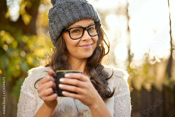 Obraz beautiful woman in a cap and glasses holds in her hand a cup of coffee in an autumn forest