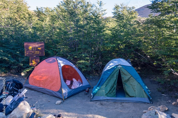 Fototapeta Two opened and messy camping tents and chaotic gear at a remote campsite in the wilderness , next to a forest in the morning or the afternoon, with a ilegible sign
