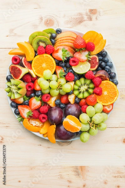 Fototapeta Healthy fruit platter, strawberries raspberries oranges plums apples kiwis grapes blueberries on the light wooden pine table, top view, copy space for text, selective focus