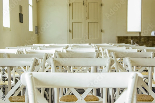 Fototapeta Partial view of white French cross-back timber chairs arranged for an event inside an old building. Seating is set up for Wedding Ceremony. No people. 