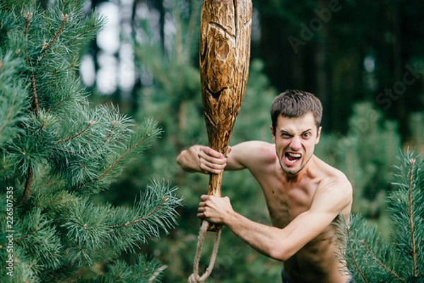 Obraz Odd primitive naked man with huge wooden stick hunting in forest. Adult male have fun like crazy psyche with rude club in hands. Expressive excited boy face.  Wild masculine strength. Cruel warrior.