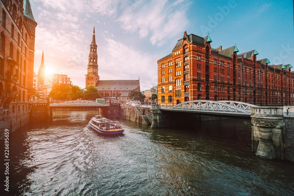 Fototapeta Turystyczny rejs statkiem po kanale z mostami w starej dzielnicy magazynowej Speicherstadt w Hamburgu w złotej godzinie zachodzącego słońca, Niemcy
