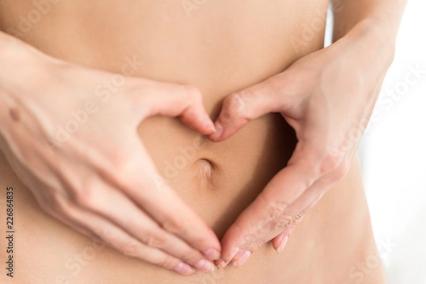 Fototapeta Caucasian woman without clothes holds fingers in the form of a figure a heart before a stomach. Light background