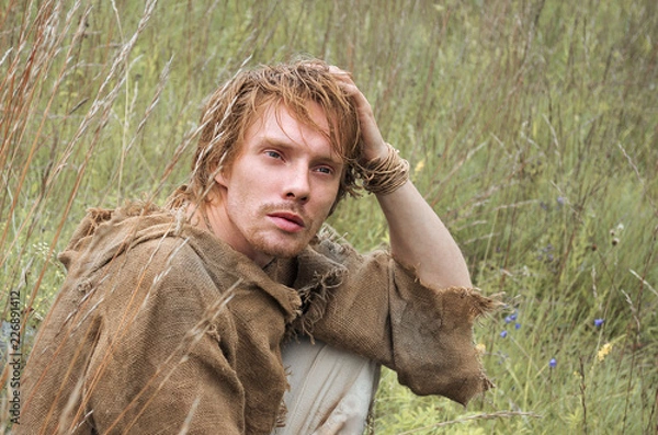 Fototapeta young shaggy and unshaven guy dressed as a medieval peasant from an old bag thought about something sitting in a field among the yellowed wild herbs