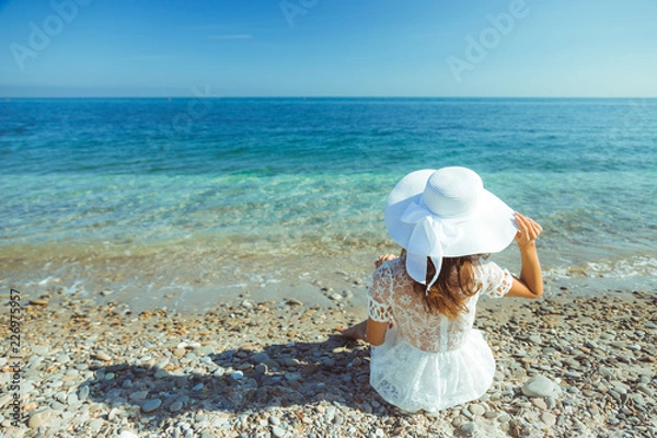 Obraz The back view of young beautiful woman in white dress sitting on the beach and looking at the sea horizon, image with copy space - concept of traveling and luxury vacation In Italy - shot from behind