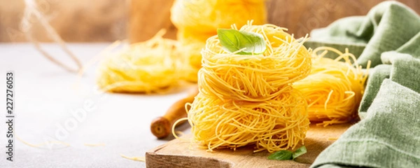Fototapeta Fresh egg pasta tagliatelle nest on wooden cutting board with basil leaf. Healthy italian food concept with copy space. Banner.