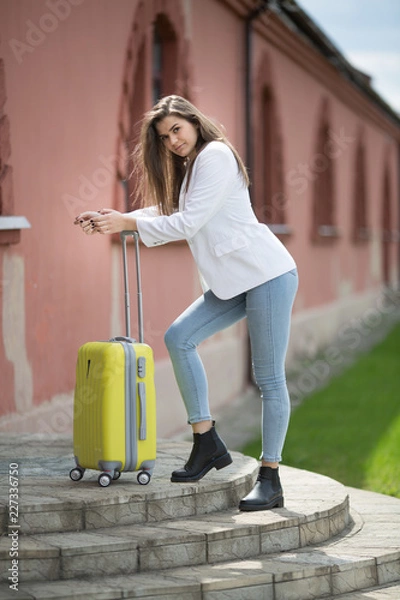 Fototapeta beautiful girl in a white jacket stands with a large yellow suitcase
