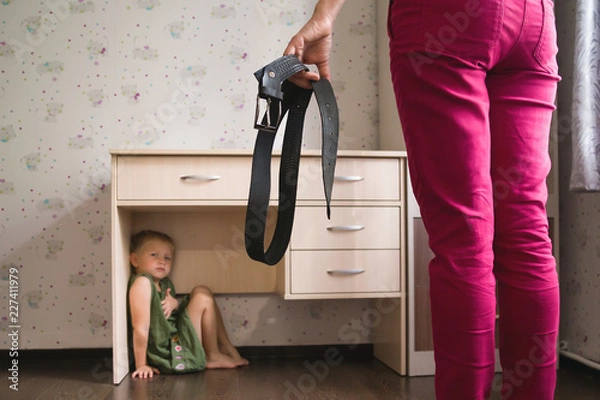 Obraz Domestic violence. Mother holding the belt in her hand to punish her small daughter. Scared child hiding under a table and sitting there terrified by physical punishment