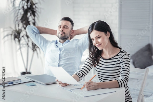 Fototapeta Time to relax. Charming girl expressing positivity while looking at documents and keeping smile on her face while making notes