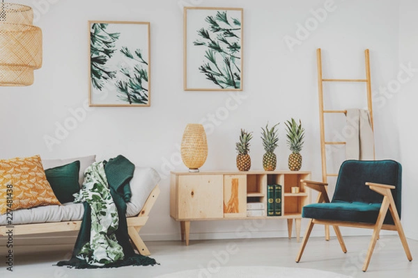 Fototapeta White living room interior with retro armchair, wooden cupboard with books, straw lamp and three pineapples, posters on wall and couch with blankets in the real photo
