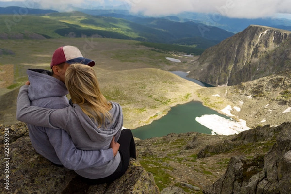 Fototapeta a loving couple sitting on the edge of a rock embraces in front of them a picturesque view of the mountains, a valley of turquoise lakes with snow and a blue sky with white clouds. Rear view follow me