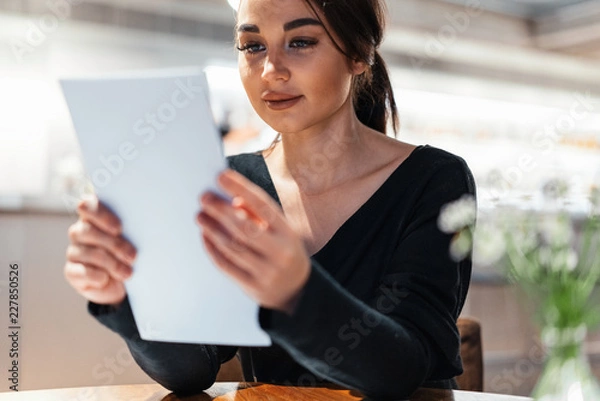Obraz Young beautiful woman looking at menu deciding what to order in modern cafe.