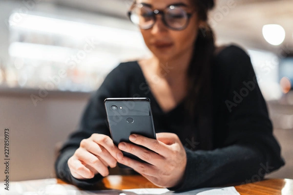 Obraz Young beautiful woman holding mobile phone in hands while waiting for her meal.