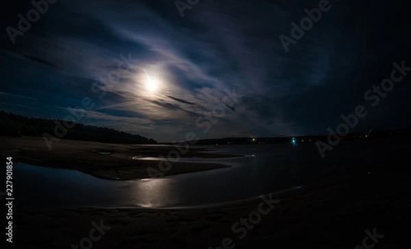 Obraz A long exposure shot of Tom river with moonlit water and shores under the cloudy night sky, foot-marks on sand.