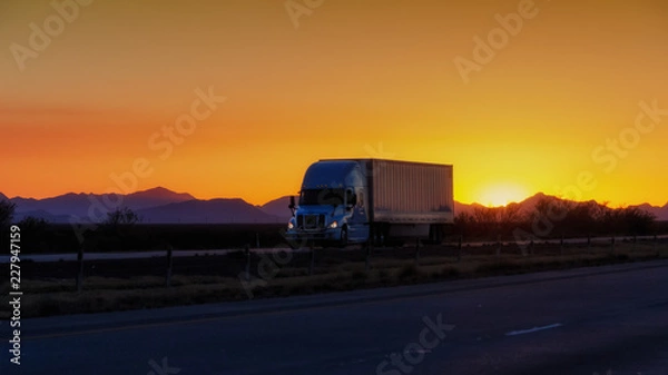 Obraz Semi-truck driving on highway at sunset with mountains in background