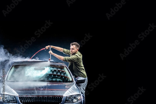 Fototapeta A man with a beard or car washer washes a gray car with a high-pressure washer at night in on the street