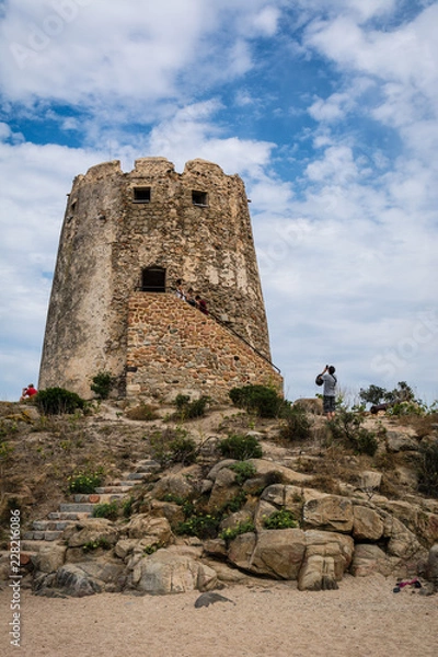 Obraz Old watchtower on the beach of Torre di Bari, Sardinia, Italy. Beautiful blue sky with clouds in the background, a path visible and some unrecognizable people.