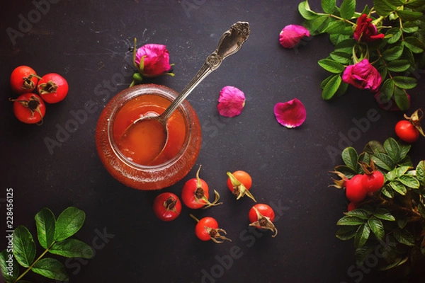 Fototapeta Spoon in a jar of rosehip jam and its berries with leaves nearby, top view