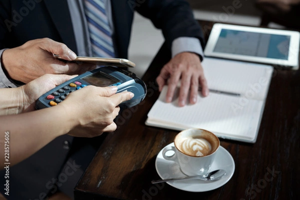 Fototapeta Businessman holding smartphone against terminal to pay for order in coffeeshop