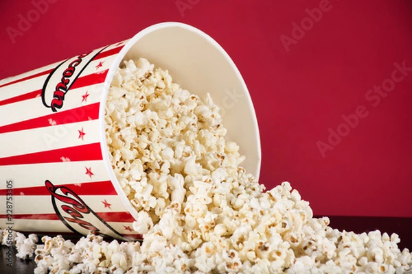 Fototapeta Spilled popcorn box on black desk and red background. Retro white and red box. Cinema and movie food concept. Close up, selective focus