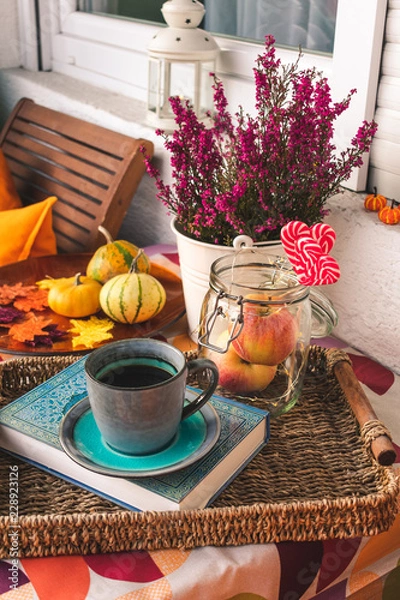 Fototapeta View of autumn balcony with pumpkins, leaves, purple calluna, apples, coffee in a vintage cup on a book and orange cushions. Autumn balcony design and decorations