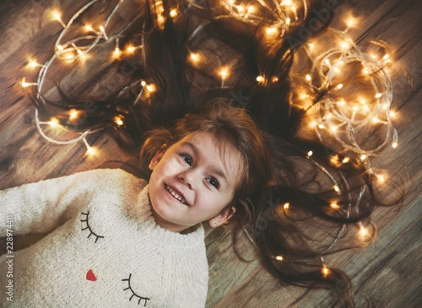 Obraz Śliczna mała dziewczynka na podłodze z jasną girlandą świąteczną we włosach.