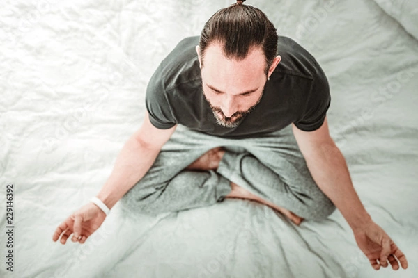 Obraz Peace of mind. Top view of a nice handsome man meditating while sitting on his bed