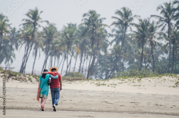 Fototapeta Lovers - man and girl go along the beach on the background of palm trees