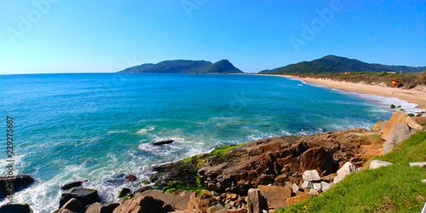 Fototapeta Caldeirão and Armação beaches in Florianopolis, Brazil, during a sunny morning. Rocks can be seen close to the photographer, and mountains can be seen far away.