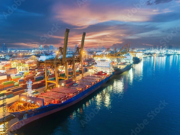 Fototapeta Container ship from sea port warehouse with working crane bridge unloading containers shipment at dusk. Suitable use for transport or import export to global logistics concept.