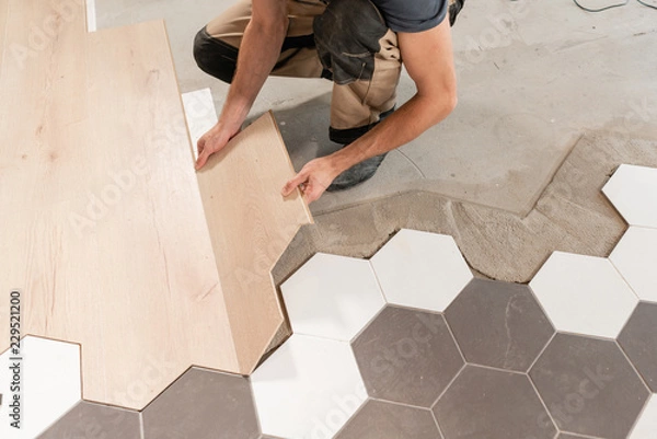 Fototapeta Male worker installing new wooden laminate flooring. The combination of wood panels of laminate and ceramic tiles in the form of honeycomb. Kitchen renovation.