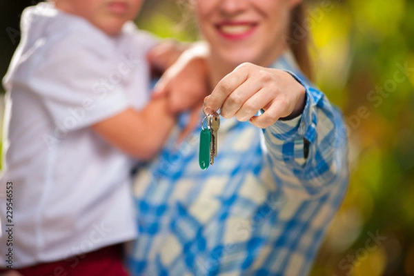 Fototapeta Young positive mother holds a small child in arms and the keys to a new apartment or house standing outside on a sunny summer warm day. Mortgage and housewarming concept