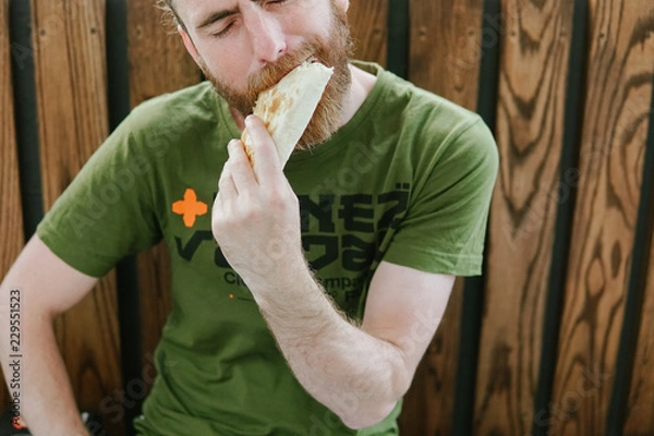 Fototapeta Street food. hipster man with a beard on the street on the bench eating doner kebab. wooden background 