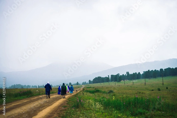 Fototapeta People go forward in mountain despite bad weather. Travelers with dog goes upwards along road. Way on foot in highlands in rainy overcast day. Summit despite everything. Walking in mountains.
