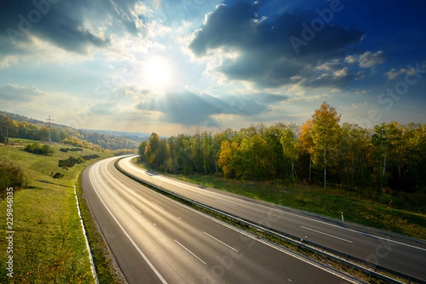 Obraz Empty asphalt highway between deciduous forest in autumn colors under the radiant sun and dramatic clouds. View from above.