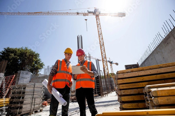 Fototapeta Civil architect  and construction manager  dressed  in orange work vests and  helmets discuss  a building project on the mobile tablet on the open building site next to the crane