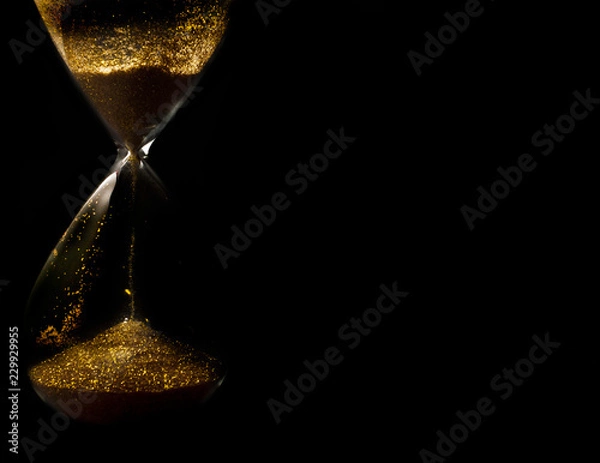 Fototapeta Sand and golden glitter passing through the glass bulbs of an hourglass measuring the passing time as it counts down to a deadline or closure on a black background with copy space