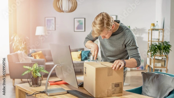 Fototapeta Handsome Young Man in Living Room with Cardboard Box Package, Opening it With Interest, Using Knife.