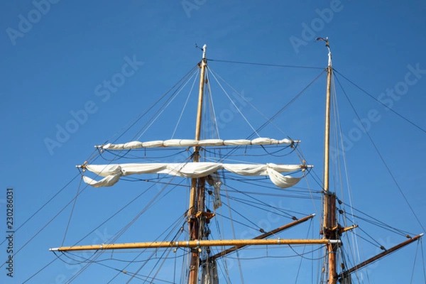 Obraz The furled sails and rigging of a 3-masted schooner, seen against the backdrop of a bright blue cloudless morning sky, while moored at an inlet in Kökar, Åland Islands, Finland.