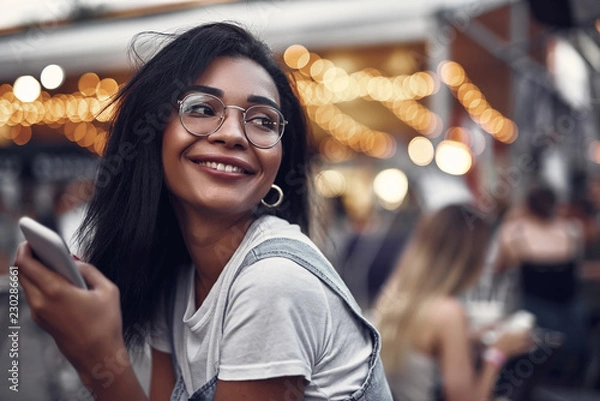 Fototapeta Portret młodej damy hipster w okularach, trzymając smartfon i odwracając się z uśmiechem. Ulica z ludźmi na niewyraźne tło z efektem bokeh