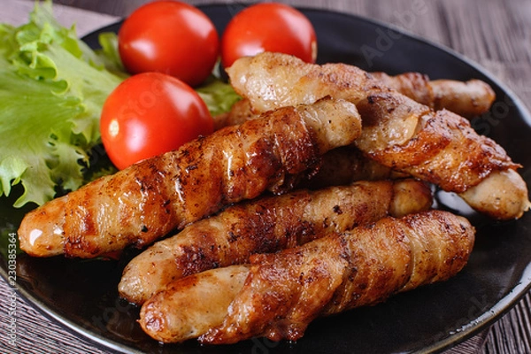 Fototapeta Appetizing and colorful sausages wrapped in bacon and grilled next to tomatoes and lettuce are on a wooden table
