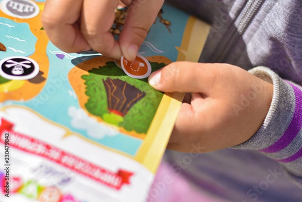 Fototapeta A child plays a pirate quest on a paper map, hands glue a sticker