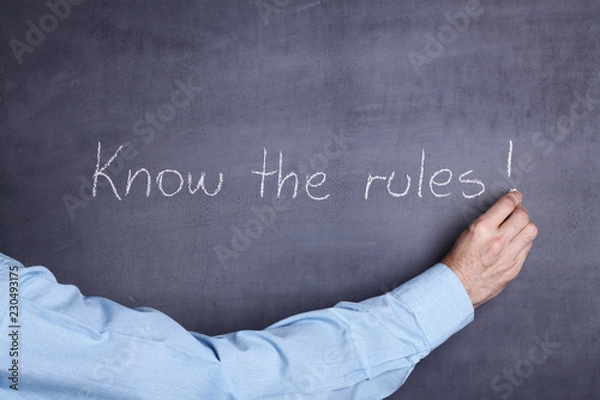 Fototapeta A man's hand holding a piece of chalk and writing the phrase: "Know the rules!" on a blackboard