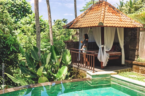 Fototapeta A young woman with a glass of juice in her hands is standing by the private pool against the backdrop of a romantic gazebo and tropical plants. A girl enjoys relaxing at a villa in Bali in Ubud.