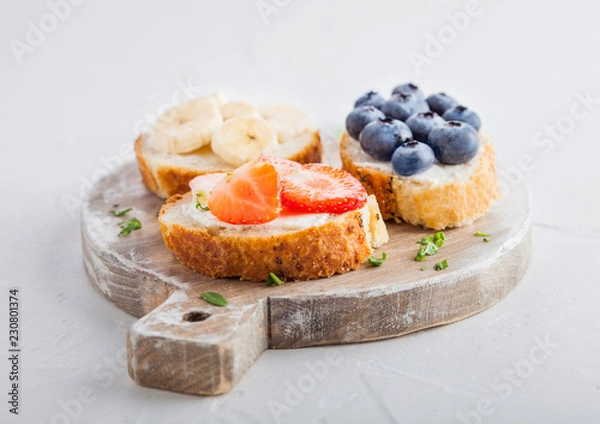 Obraz Fresh healthy mini sandwiches with cream cheese, fruits and berries on vintage wooden board. Strawberries, blueberries, bananas and raspberries on stone kitchen table background.Top view.