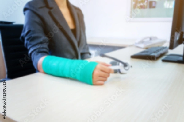 Fototapeta blurred woman with broken hand and green cast  working on computer in office