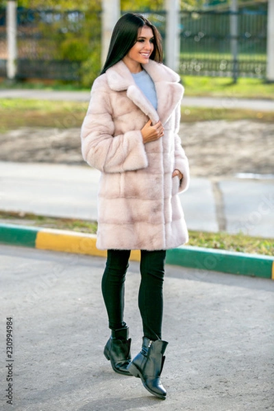 Obraz Beautiful sexy brunette in a light fur coat and black pants walks down the street and smiles